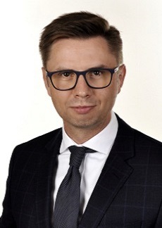 Mężczyzna w ciemnym garniturze, białej koszuli i ciemnym krawacie. Ma ciemne włosy i nosi okulary. Jest to Prezes Zarządu Piotr Petelewicz